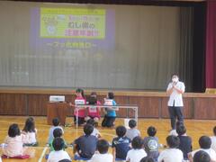体育館のステージ前で作業する3人の子供と話をする1人の大人を見ている複数の子供たちの写真