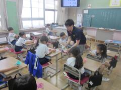 教室で、体操着姿の女の子に話しをする男性を見ている子供たちの写真