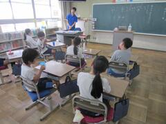 教室の席に着く子供たちと前に立つ青い服の大人の写真