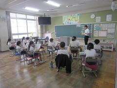 教室の席に着き黒板前に立つ大人の話を聞く子供たちの写真