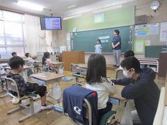 テレビの点いた教室の前に立つ子供を見ている複数の子供たちと、大人に指導を受けている1人の子供の写真