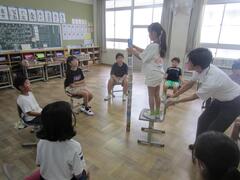 大人に抑えられた椅子に立ち空き缶を立てる子供を円になって見ている子供たちの写真