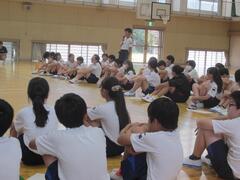 大勢の子供たちが体育館の床に座っている中で、1人が立っている写真