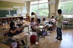 教室の席に着いて前に立つ大人を見ながら作業をする子供たちの写真