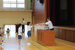 体育館のステージ前に置かれた講壇に立つ男性から資料を受け取る子供の写真