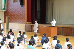 体育館のステージ前に置かれた講壇に立つ大人とその横に立つ子供を見ている子供たちの写真