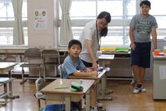 席に着いてこちらを見ている男の子に話しかけている大人とその横に立つ大人の写真
