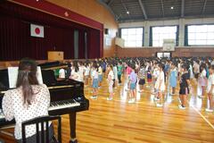 体育館で、手前にはピアノとそれを引く大人の女性が、その向こうに大勢の子どもたちが立っている写真