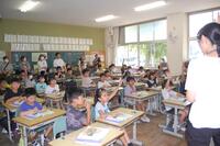 教室で何人か子どもたちが手を挙げていて、手前には大人の女性が写っている写真