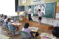 教室の教壇に大人と子供がひとりずつ立、座っている複数の子どもたちがそれを見ている写真