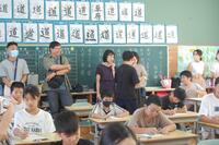 教室の後ろ側に幾人かの大人たちがたっていて、書き物をしている子どもたちを眺めている写真
