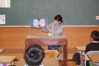 教室の教卓の椅子に座って生徒たちに向かって絵本を読んでいる女性の写真