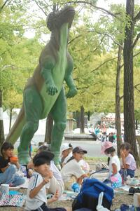 屋外の恐竜のオブジェクトの下でシートを引いてご飯を食べている生徒たちの写真