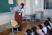 教室の黒板の前に立って絵本を読んでいる女性とそれを見上げている生徒たちの写真
