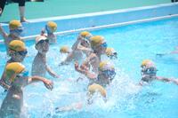 プールで黄色い水泳キャップを被った水着姿の子どもたちが水しぶきをあげながら泳いだり、立ったりしている写真