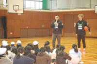体育館で、バスケットボールをわきに抱えた大人とマイクを持った大人が、床に座っている子どもたちの前で話をしている写真