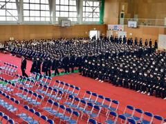 体育館で行われた入学式で黒い制服を着た大勢の入学生が両脇で座っている大勢の在校生を横切りながら入場する様子の写真