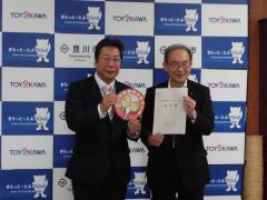 スーツ姿の男性2名がそれぞれ協定書と豊川市高齢者みまもり隊ワッペンを両手に持ち笑顔で立っている様子の写真