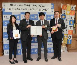 オフィスの一角で、「おもてなCITYとよかわ」と書かれた旗の前でスーツ姿の男女4人がそれぞれ協定書か豊川市高齢者みまもり隊ワッペンを持って横並びで立っている写真