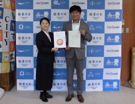 グレーのスーツを着た男性と黒いスーツを着た女性がそれぞれ協定書と豊川市高齢者みまもり隊ワッペンの印刷された書類を両手に持って横に並んで立っている写真