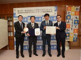 オフィスの一角で、「おもてなCITYとよかわ」と書かれた旗の前でスーツ姿の男性4人がそれぞれ協定書か豊川市高齢者みまもり隊ワッペンを持って横並びで立っている写真
