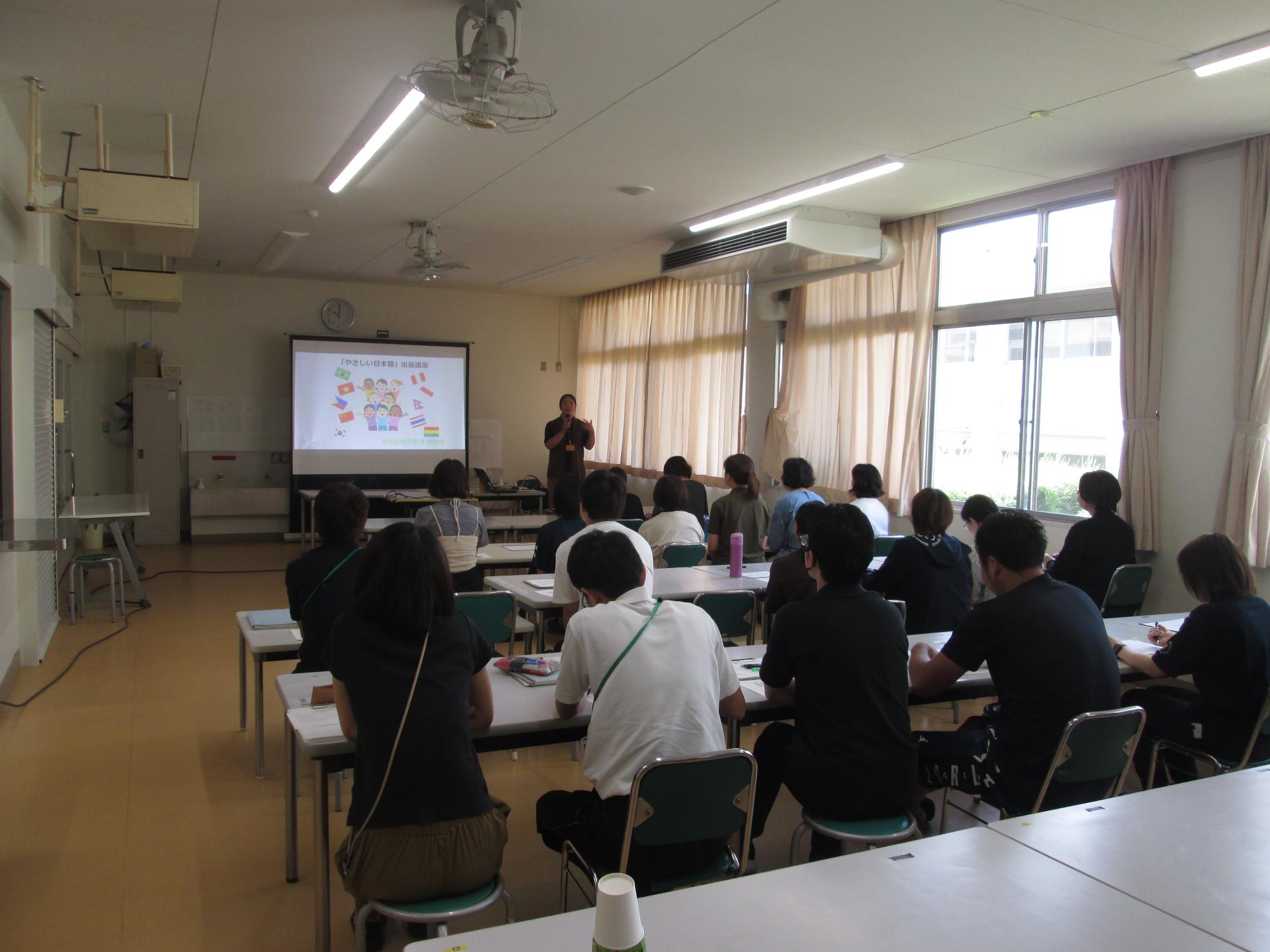 非遮光カーテンがかかった室内の椅子に座って机の上の紙に記入をしている人々の写真