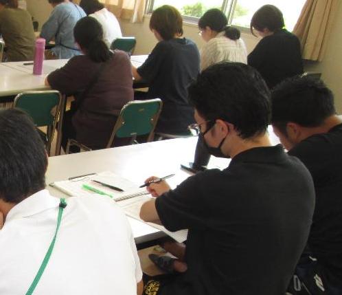 非遮光カーテンがかかった室内の椅子に座って机の上の紙に記入をしている人々の写真