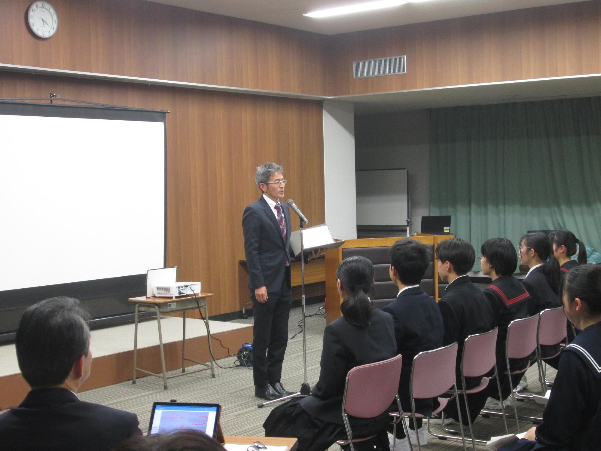 スーツを着た男性が座っている中学生や大人に対して話をしている写真