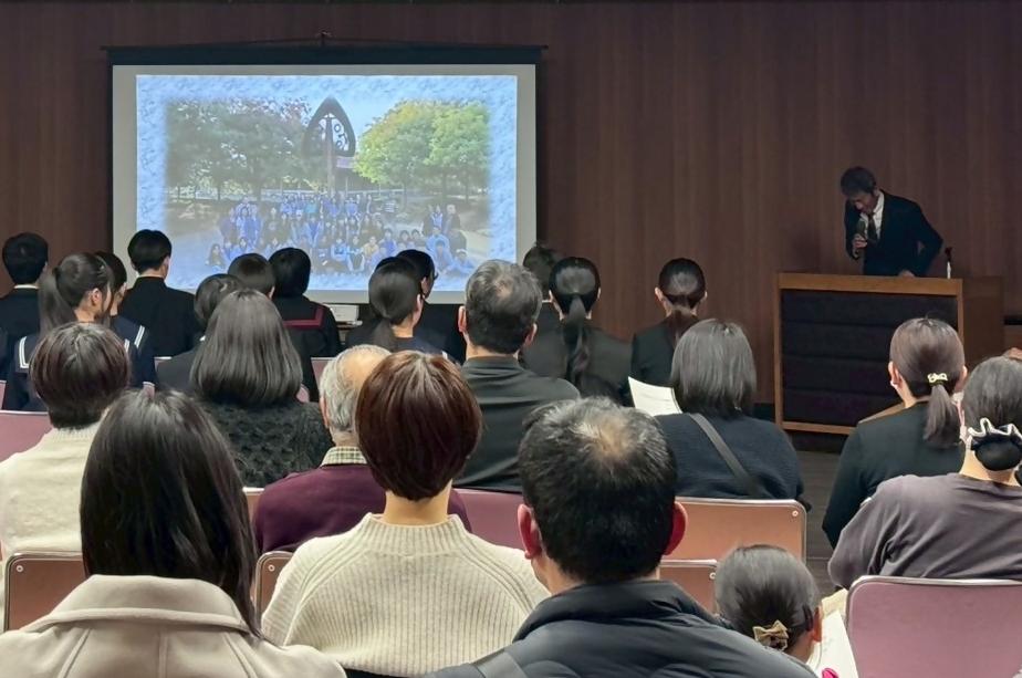 スーツを着た男性がスクリーンに映像を映しながら座っている中学生や大人に対して話をしている写真