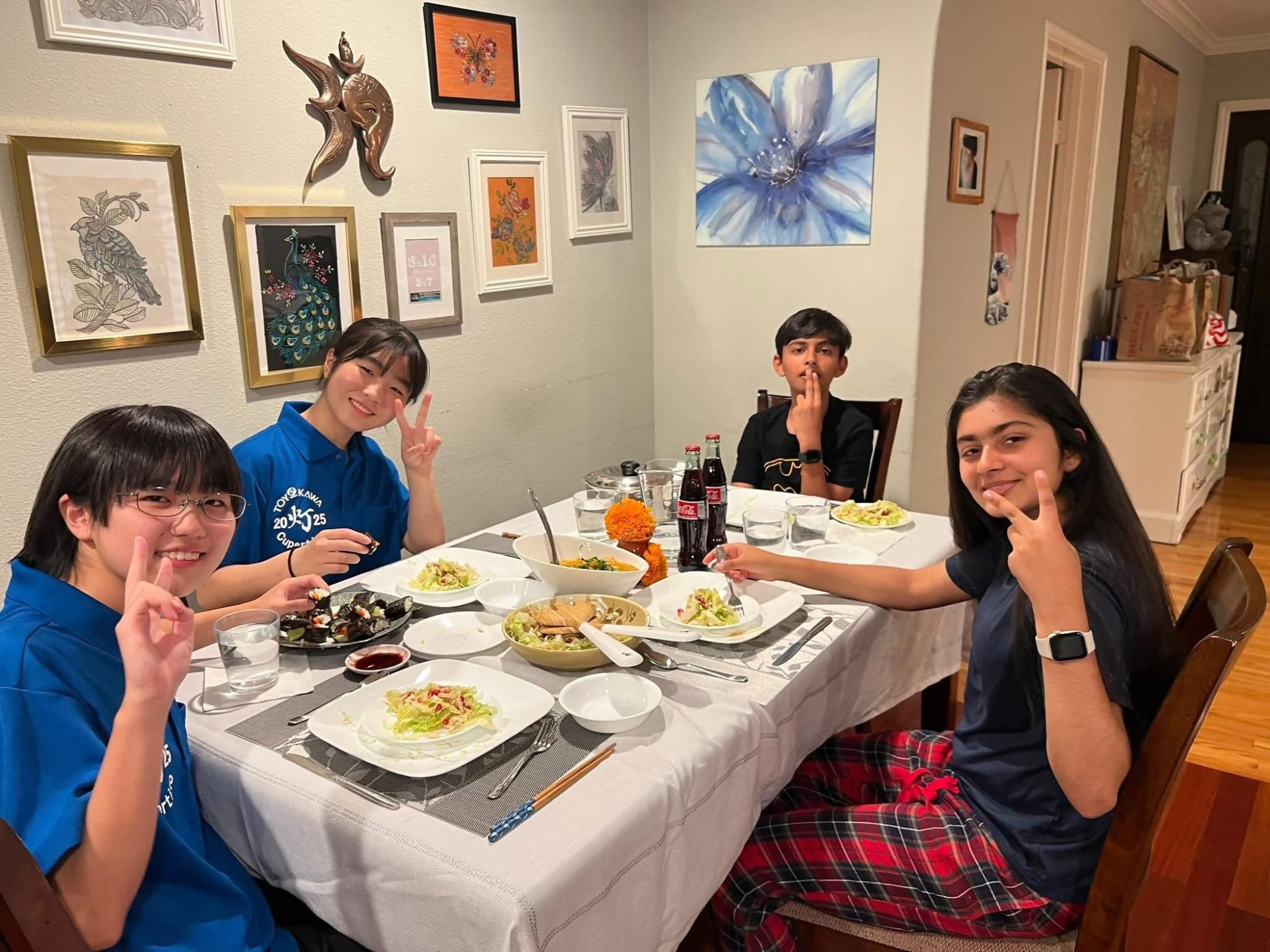 青色のポロシャツを着た日本人の女子生徒2人とアメリカ人の姉弟がテーブルで食事をしている写真