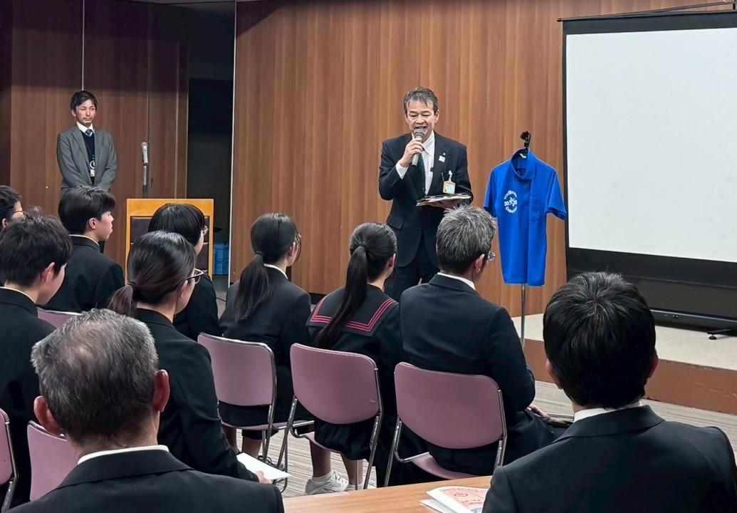 スーツを着てマイクを持った男性が中学生や大人の前で話をしている写真