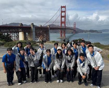 橋を背景に、灰色のパーカーや青色のポロシャツを着た使節団員の集合写真