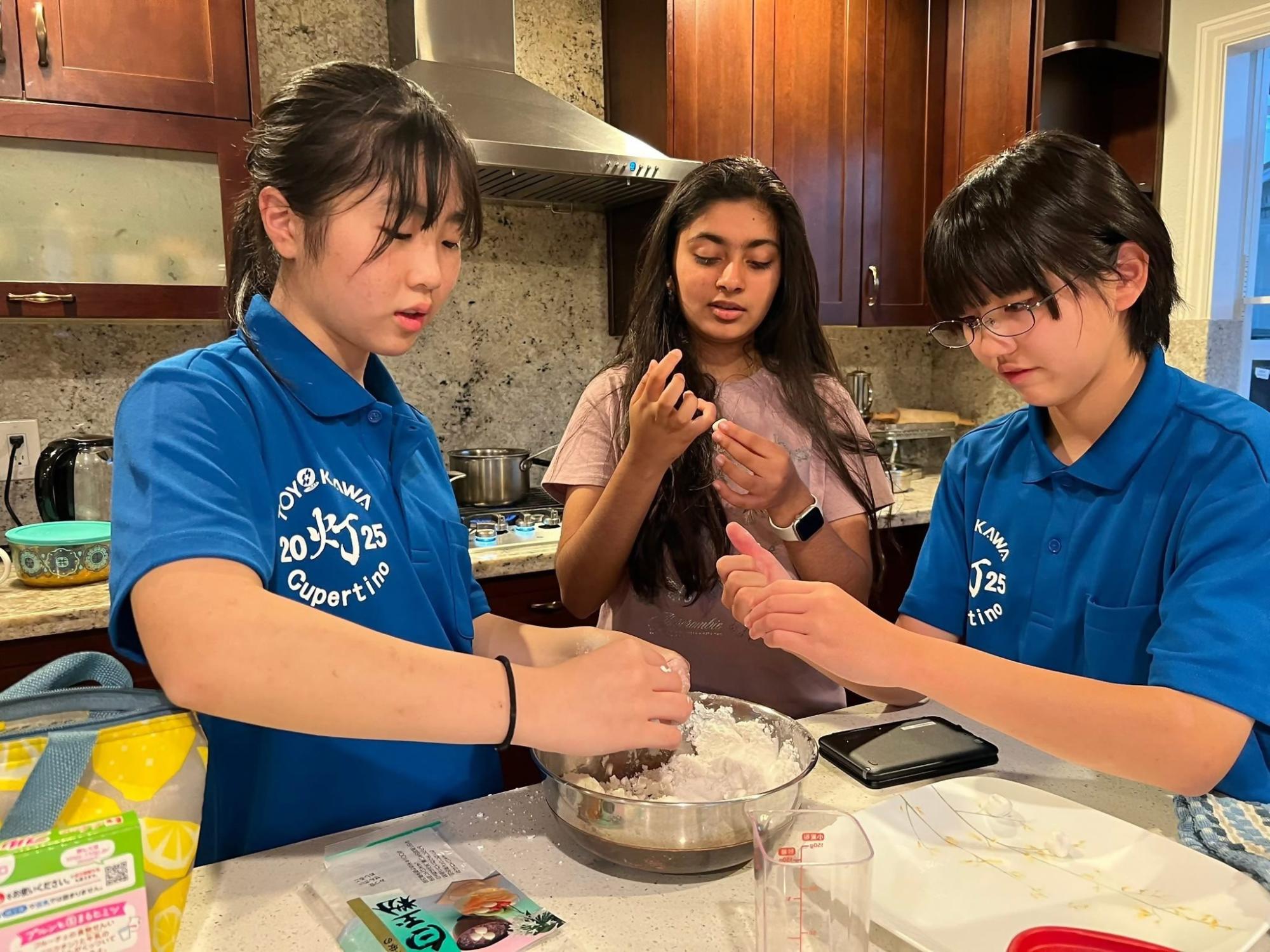 青い服を着た日本人の女の子二人とピンクの服を着たアメリカ人の女の子一人が料理をしている写真