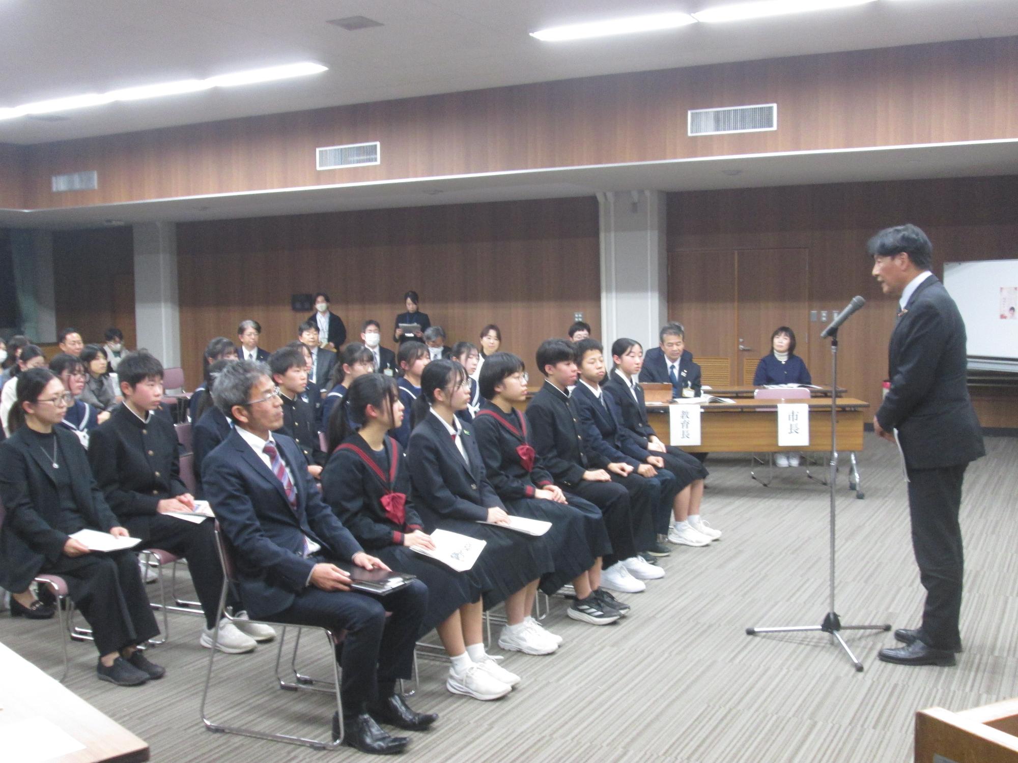 市長が座っている使節団員に向かってスタンドマイクで話をしている写真