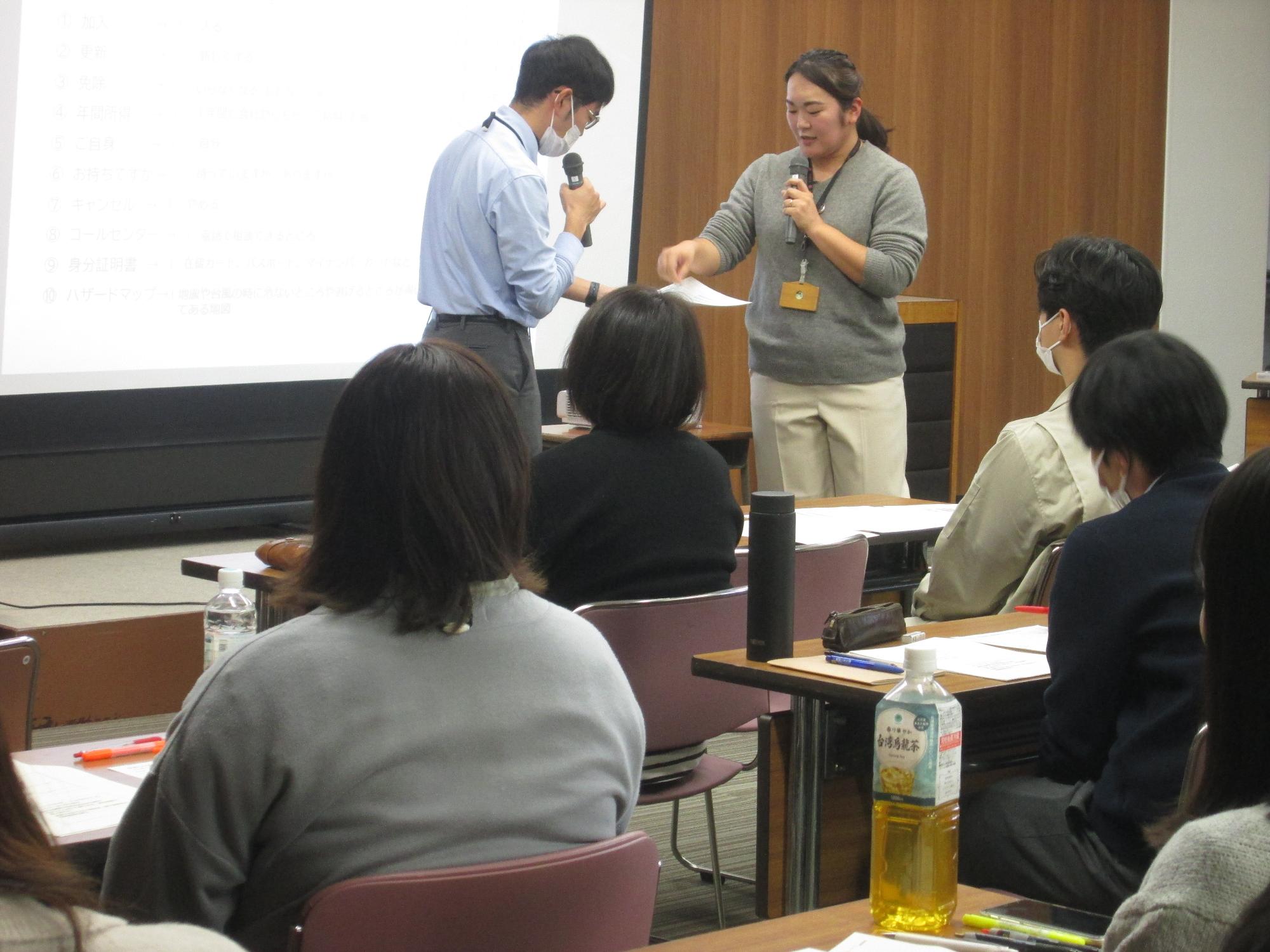 スクリーンの前に立ってマイクで話をしている2人の職員の写真