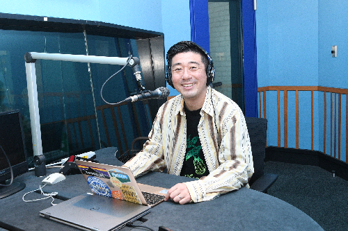 DJブースの中で、マイクの前でヘッドホンをし、パソコンを開いている笑顔の男性の写真