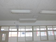 中学校の教室の天井に大型クーラーが設置されており、その下に扇風機が壁に取り付けられている写真