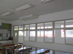 中学校の教室の天井に大型クーラーが設置されており、その下に扇風機が壁に取り付けられている様子を別角度から写した写真