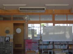 小学校の教室の天井に大型クーラーが設置されており、その下に扇風機が壁に取り付けられている写真