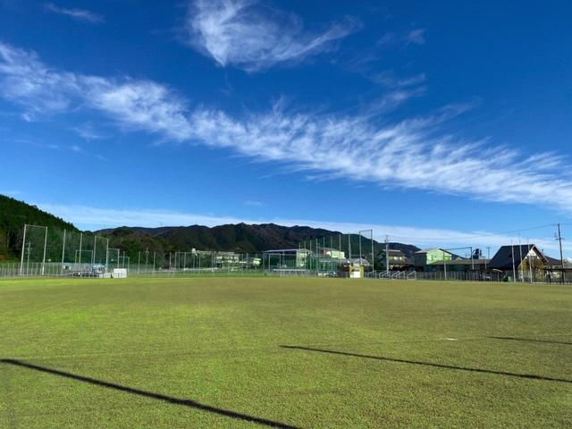 きれいな青空の下、緑豊かな山々が奥に見え、緑色のネットが設置されているスポーツ公園サッカー場の芝グラウンドの写真