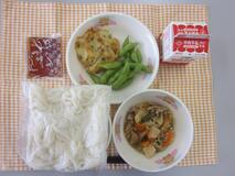テーブルクロスに並べられた冷やし白玉うどん・牛乳・ぶた肉とやさいの炒め煮・かき揚げ・えだ豆・うどんつゆの写真