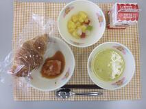 テーブルクロスに並べられたクロワッサン・牛乳・米粉のコーンクリームスープ・とり肉のトマトソースかけ・フルーツカクテルの写真