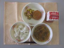 テーブルクロスに並べられた麦ごはん・牛乳・沢煮わん・和風コロッケ・ツナそぼろ丼の写真