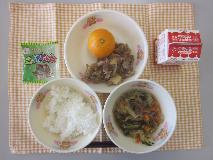 テーブルクロスに並べられたごはん・牛乳・はるさめスープ・ぶた肉のしょうが炒め・みかん・かんそう小魚の写真