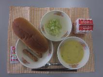 テーブルクロスに並べられたサンドイッチロールパン・牛乳・米粉のコーンクリームスープ・ホットドッグ（ウインナー/キャベツソテー）・ミニケチャップの写真