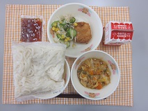 テーブルクロスに並べられた冷やし白玉うどん・牛乳・ぶた肉とやさいの炒め煮・いかメンチカツ・切り干しだいこんのサラダ・うどんつゆ・ごまドレッシングの写真