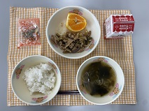 テーブルクロスに並べられたごはん・牛乳・わかめスープ・ピリ辛丼・はるみみかん・アーモンド小魚の写真