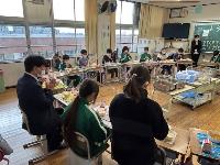 円状に並べられた机の上に給食が乗せられ、椅子に市長と児童が座っている写真