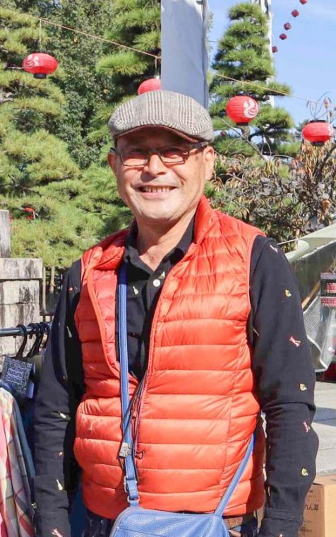 神社の境内のような場所に複数の出店が並んでいる中、カバンをぶら下げた1人の男性が微笑んで立っている写真