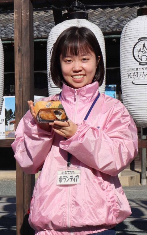 複数の提灯がぶら下がっている場所をバックに1人の女性がパック容器に入ったいなり寿司を手に持ち微笑んでいる写真